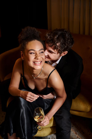 handsome man in suit embracing joyful african american woman with glass of wine and sitting on sofaの写真素材