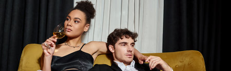 elegant african american woman drinking wine and sitting with man in suit on velvet sofa, banner