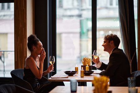 happy interracial couple in elegant attire holding glasses of wine during date in restaurant, laugh