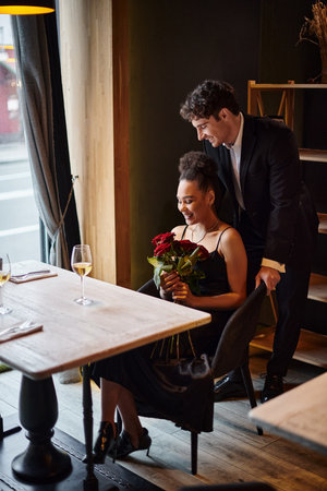 gentleman standing behind chair of happy african american woman holding roses during dateの写真素材