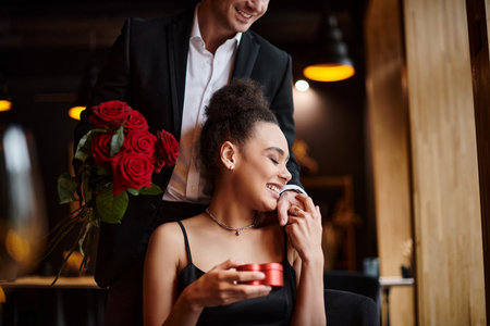 man holding red roses near happy african american woman with heart-shaped box on Valentines dayの写真素材