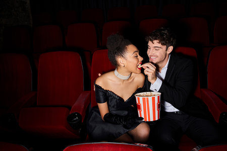 joyful handsome man in black suit feeding popcorn to his african american young girlfriend on dateの写真素材
