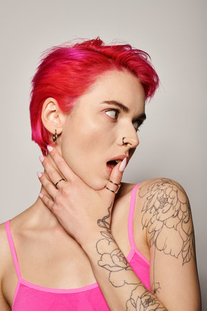 tattooed and pierced woman with pink hair and rings on her fingers looking away on grey backgroundの写真素材