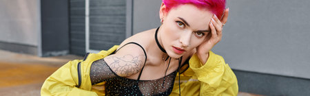 alluring stylish woman with pink hair and tattoos posing on street and looking at camera, banner