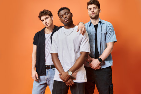appealing multiracial male models in street attires looking at camera on orange backdrop, fashion