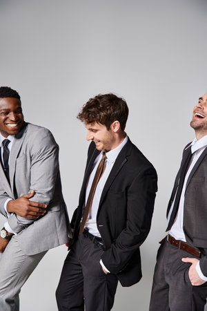 attractive cheerful multicultural male models in smart suits smiling sincerely on gray backdropの写真素材