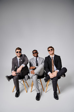 cheerful young multicultural men in business smart suits sitting on chairs and smiling happilyの写真素材