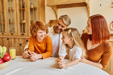 joyful parents with daughter and son sitting near fresh fruits and orange juice in cozy kitchen