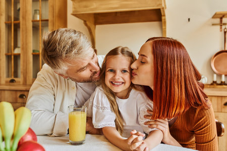 caring parents kissing daughter near fresh fruits and orange juice in kitchen, love and careの写真素材
