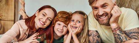 joyful parents with son and daughter lying ibn cozy bedroom and looking at camera, horizontal bannerの写真素材
