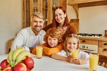 joyful couple with adorable kids looking at camera near fresh fruits and orange juice in kitchen