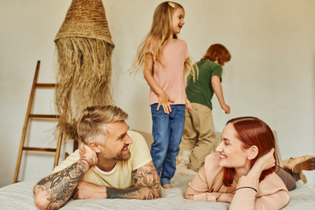 husband and wife smiling at each other near happy kids having fun on bed at home, relaxation timeの写真素材