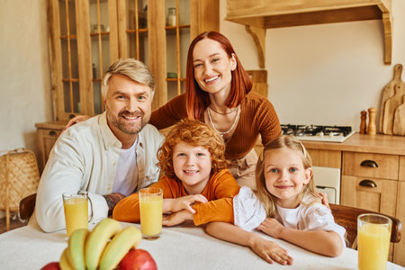 joyous parents with adorable kids looking at camera near fresh fruits and orange juice in kitchenの写真素材