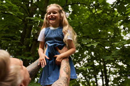overjoyed girl laughing in hands of tattooed father in green park, leisure and playing together