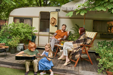 kids learning to play acoustic guitar while parents talking in deck chairs near trailer homeの写真素材