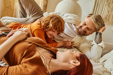 happy tattooed man looking at wife and redhead son having fun on bed at home, relaxation timeの写真素材