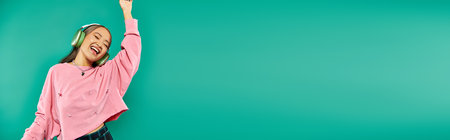 positive young asian girl in wireless headphones gesturing and dancing on turquoise, banner