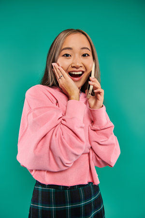 portrait of amazed young asian woman in pink sweatshirt talking on smartphone on turquoise backdropの写真素材