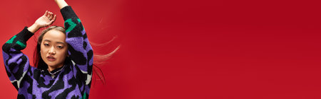 pretty young asian girl in vibrant sweater with animal print posing with raised hands on red, bannerの写真素材