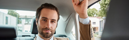 successful and handsome businessman in beige elegant suit looking at camera in luxury car, banner