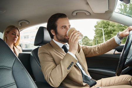 stylish man in formal wear drinking coffee to and driving car with blonde businesswoman on rear seat