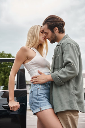 trendy bearded man embracing sensual blonde woman near car on modern urban street, affectionの写真素材