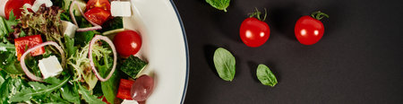 top view photo of plate with traditional Greek salad near cherry tomatoes on black backdrop, banner