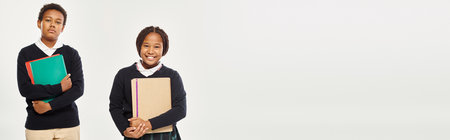 happy african american schoolkids in neat uniform holding textbooks and standing on grey, banner