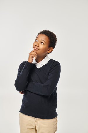 pensive african american boy in school uniform touching chin while thinking on grey backdropの写真素材
