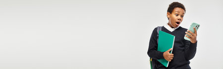 shocked african american schoolboy in uniform holding smartphone and notebook on grey, bannerの写真素材