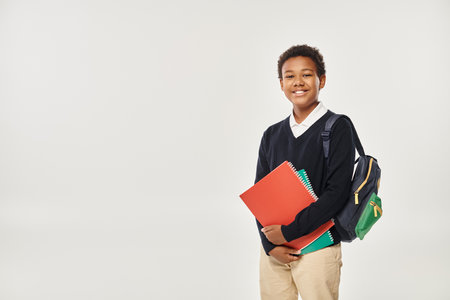 happy african american schoolboy in uniform holding notebooks and standing on grey backgroundの写真素材