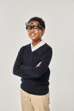 cheerful african american boy in sweater and eyewear holding glasses and looking at camera on grey