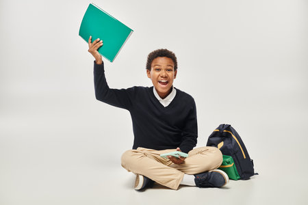 happy african american boy in uniform holding smartphone and notebook while sitting near backpackの写真素材