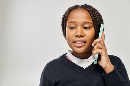 preteen african american schoolgirl in uniform talking on smartphone on grey background, look down