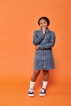 full length of cheerful african american girl in denim dress smiling on orange backgroundの写真素材