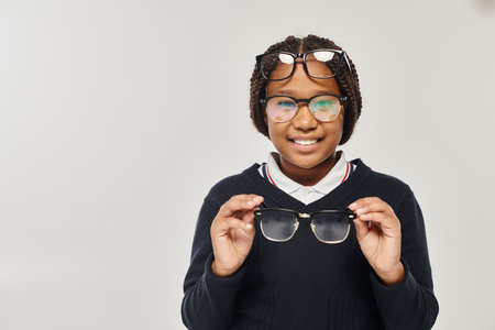happy african american girl in sweater and eyewear holding glasses and looking at camera on greyの写真素材