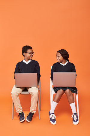 happy african american kids in uniform sitting on chairs and using laptops on orange backdrop