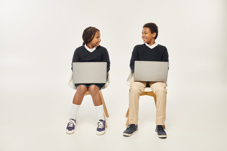 cheerful african american schoolkids in uniform using laptops and sitting on grey backdropの写真素材