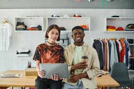 joyful multiethnic designers with laptop and coffee to go looking at camera in textile print studio