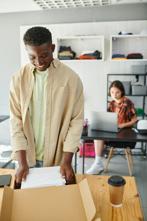 pleased african american man packing clothes in carton box near asian designer at laptop in atelier