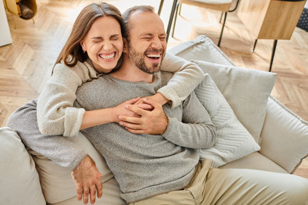 excited woman with closed eyes embracing husband laughing on couch in living room, child-free couple