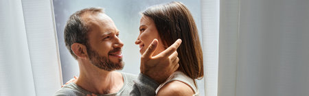 tender husband touching hair of smiling wife near window at home, child free couple, banner