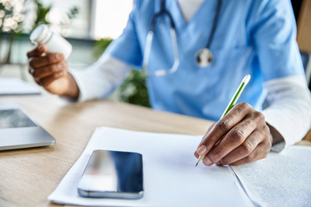 cropped view of african american doctor with stethoscope taking notes while working, phone on tableの写真素材