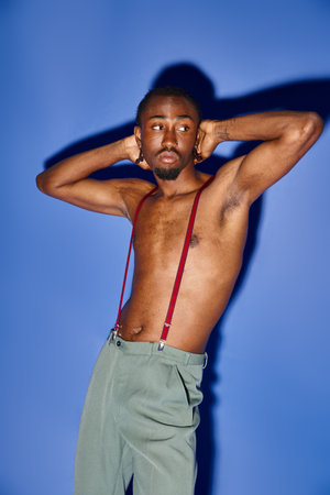 hot african american man with suspenders posing on blue backdrop and looking away, fashion conceptの写真素材