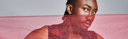 handsome african american male model posing with vivid red tulle fabric on gray backdrop, bannerの写真素材