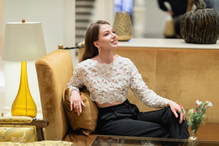 Smiling and fair haired woman in lace top and pants sitting on couch in hotel lobby of Berlin