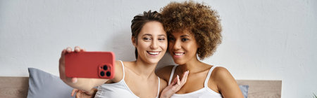 happy woman taking selfie on smartphone near african american girlfriend in bedroom, bannerの写真素材