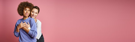 happy woman hugging her african american girlfriend on pink background, lgbtq couple love bannerの写真素材