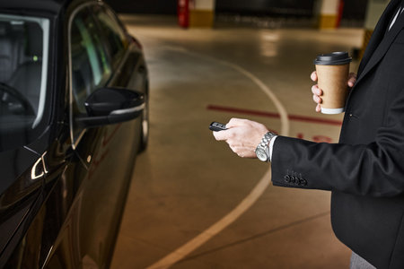 cropped view of elegant man in black suit with coffee cup in hand using car keys, business conceptの写真素材
