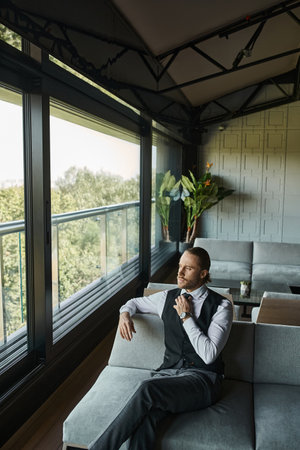 elegant businessman in black vest fixing his tie while sitting on sofa and looking out of windowの写真素材